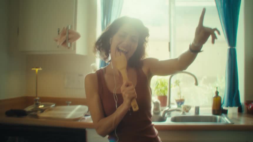 A woman dancing and singing in her kitchen while listening to music on earbuds