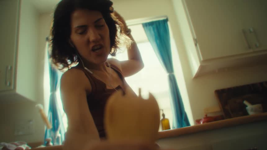 A smiling woman dances playfully with earbuds in and a wooden spoon in hand lit by warm sunlight
