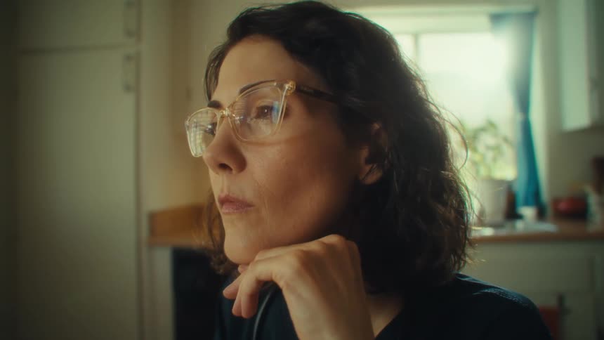 A doctor sitting in a kitchen at midday wearing a black uniform with a stethoscope over her shoulders and glasses while reading a magazine