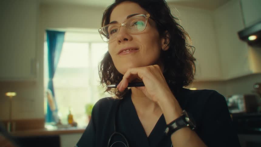 A woman in scrubs and glasses reflects thoughtfully at a kitchen table her stethoscope resting on her chest