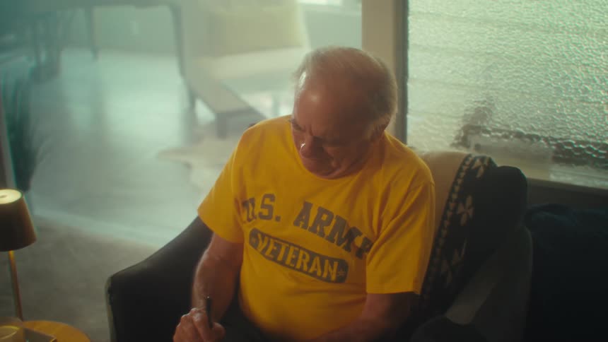 A closeup shot of an elderly man with a yellow us army veteran shirt sitting in an armchair smiling and concentrating as he writes in a notebook with a pen