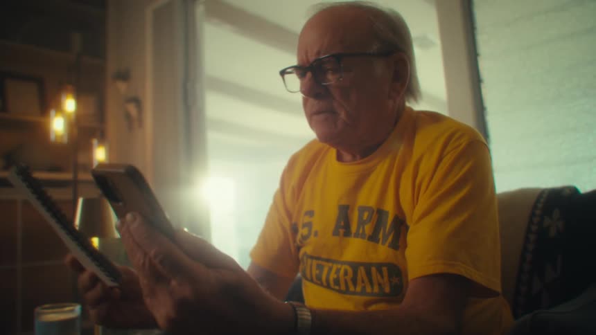 Man wearing glasses and a yellow shirt holds a phone and a notebook