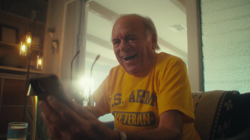 An elderly man laughs and celebrates after viewing media content on a mobile phone while sitting in a living room during daytime
