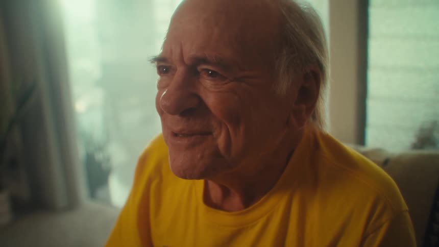 An elderly man wearing a yellow veteran tshirt sits on a couch with a smile on his face writing something down