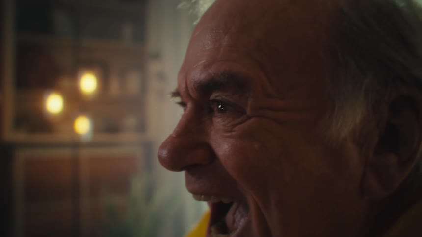 An elderly man beams with joy his face lit by sunlight as he smiles warmly indoors