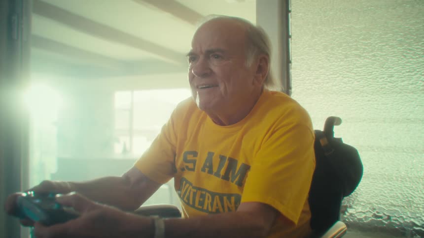 An elderly man plays video games with a controller while sitting on a wheelchair in a living room during daytime