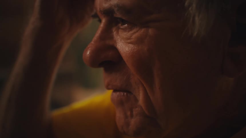 An elderly man in a yellow shirt thoughtfully holds a pen and writes in a notebook near a small round table with a glass of water