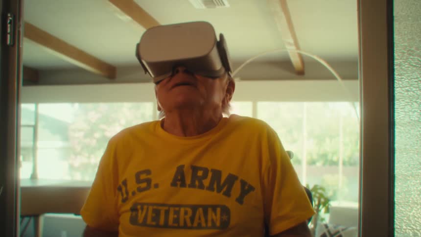An elderly man sitting in a wheelchair at midday in his living room wearing a vr headset and holding a game controller having a fun time playing a game