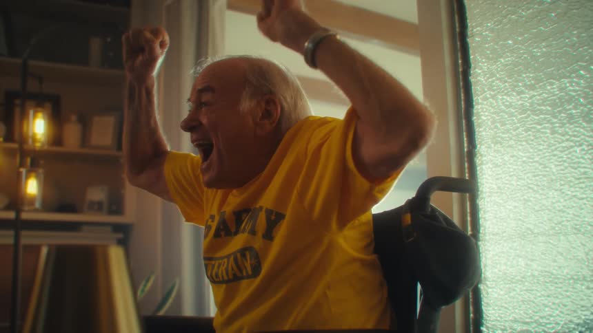 An older man in a yellow army veteran shirt cheers joyfully with both fists raised indoors