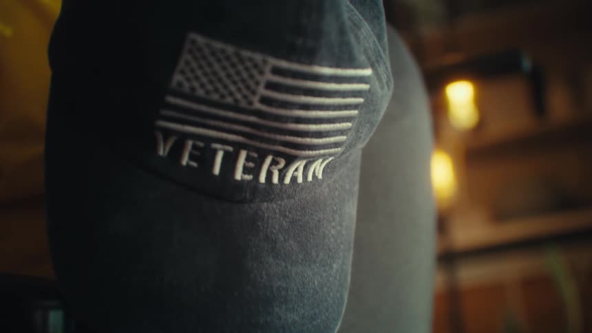 A black baseball cap with an embroidered american flag and the word veteran hangs on the back of a chair