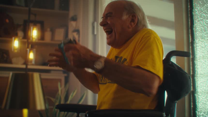 An elderly man plays a video game with a controller while sitting on a wheelchair in a living room during daytime