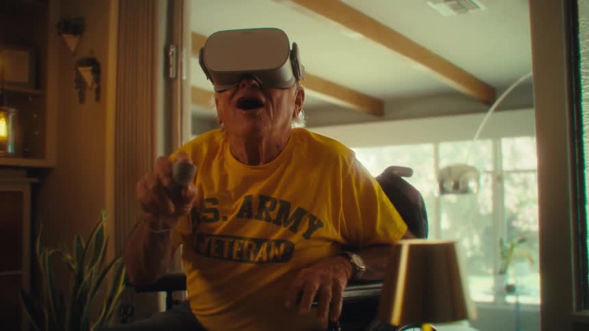 A happy elderly man in a wheelchair wearing a yellow us army veteran shirt and a vr headset smiles as he interacts with the virtual environment