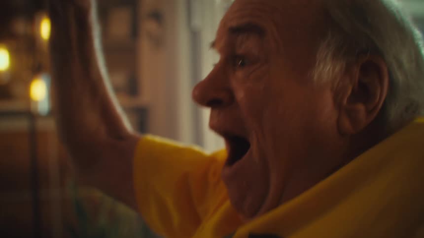 An elderly man wearing a yellow tshirt super excited while watching a game in his living room
