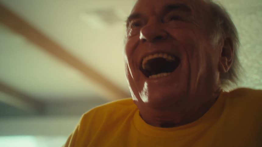 An elderly man in a yellow shirt laughs joyfully while playing with a video remote his face lit with excitement in a warmly lit room