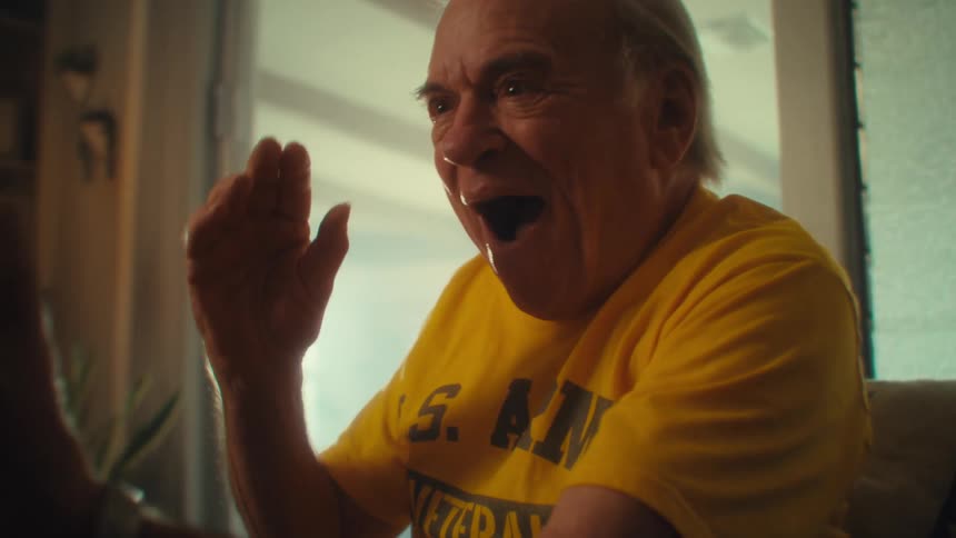 Elderly man in a yellow shirt holds his phone and speaks excitedly