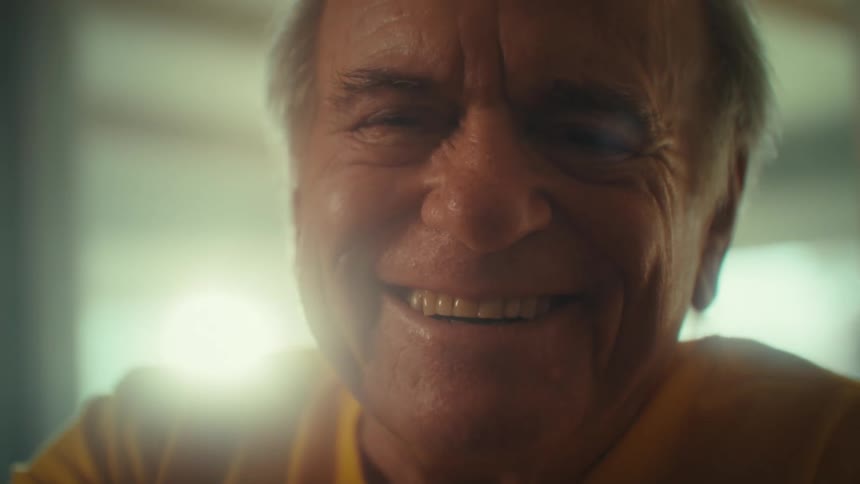An elderly man laughs as he uses a mobile phone while sitting in a living room during daytime