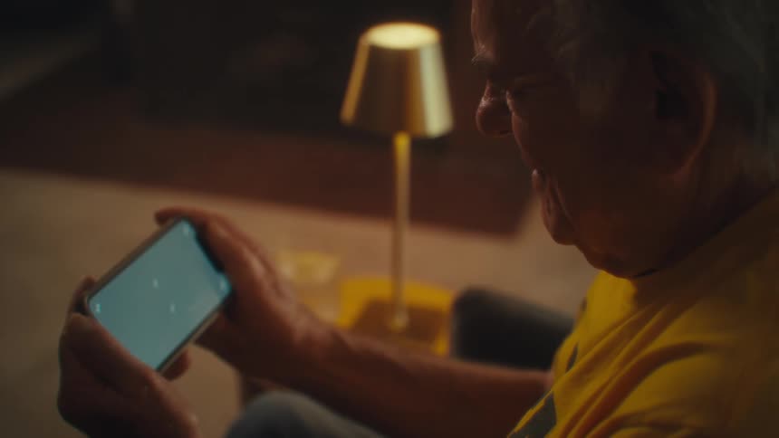 A closeup of a senior mans face and hands as he holds a smartphone horizontally and looks at it in a dimly lit room with a lamp in the background