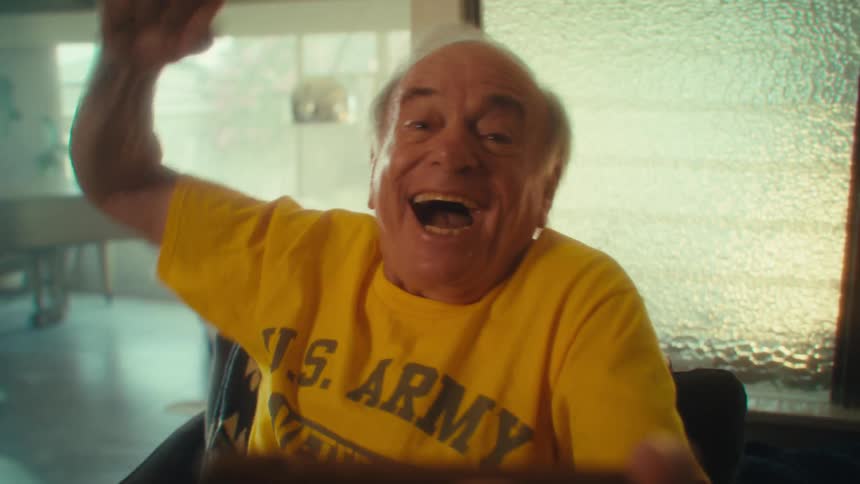 An elderly man wearing a yellow tshirt with us army veteran written on it sitting on a couch and looking super excited at the content on his phone held in his hand
