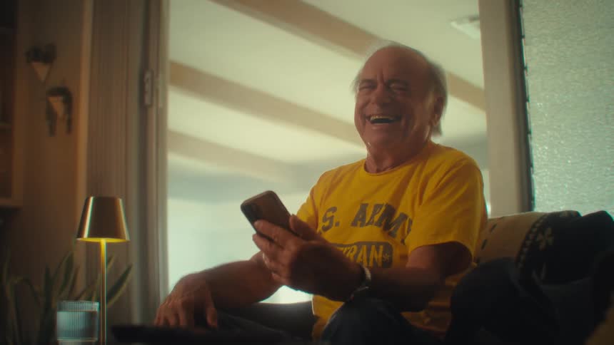 Man in a yellow shirt sitting on an armchair smiles while looking at a phone