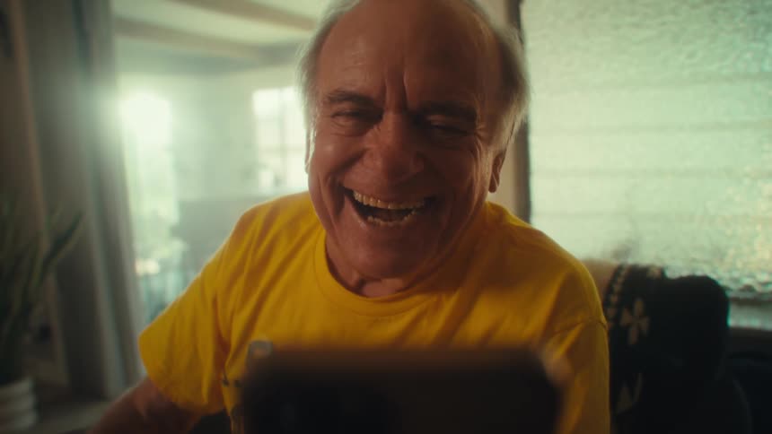 An elderly man looking overly excited during a video call on his phone sitting in a chair in his living room