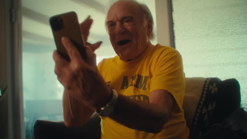 Elderly man in a yellow shirt smiles while holding his phone with both hands