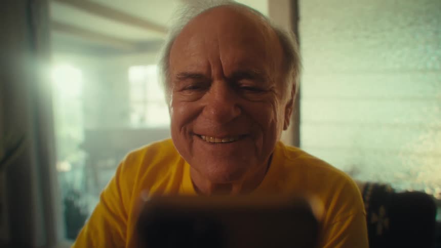 Close up portrait of an elderly man laughing while viewing media content on a mobile phone while sitting indoors during daytime