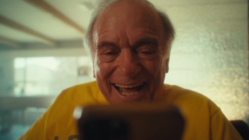 An elderly man picks up a mobile phone to take a video call while sitting in a living room during daytime
