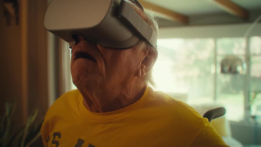 A man with white hair and a yellow shirt reacts with an expression of shock or awe while wearing a virtual reality headset and moving his head around