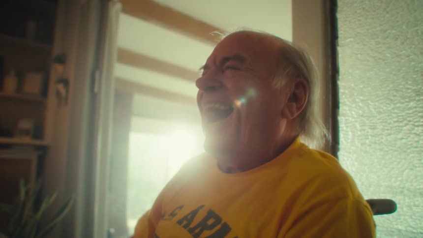 A joyful elderly man in a yellow us army shirt plays video games indoors grinning with excitement
