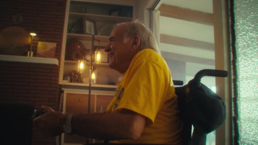 An elderly man sitting in a wheelchair in his living room holding a controller and having fun playing video games