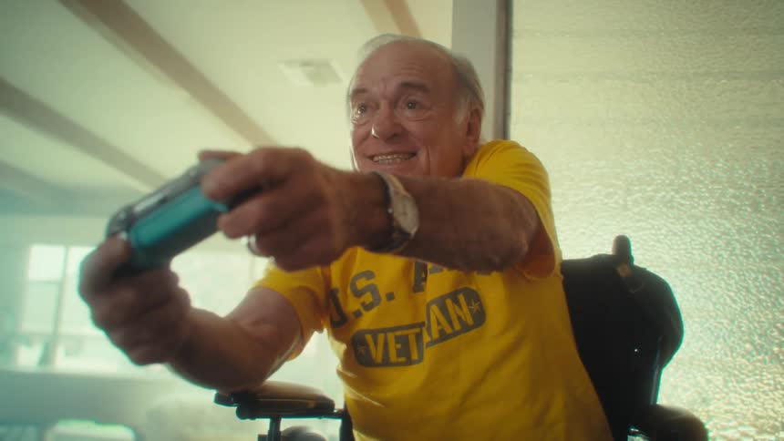 An elderly man sitting in a wheelchair playing a game with a controller having a fun time at midday in his living room