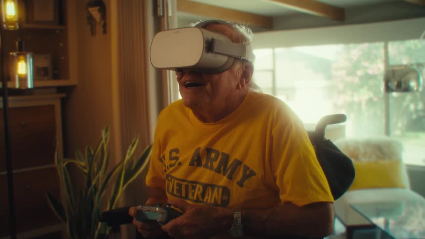 An elderly man in a wheelchair wearing a yellow us army veteran shirt joyfully uses a vr headset in a sunlit room