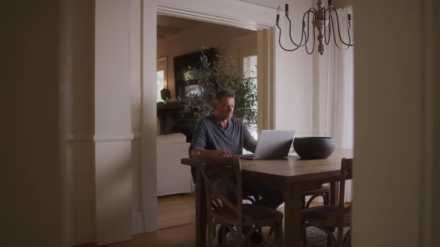 A man works at a laptop in a cozy dining room filled with soft natural light