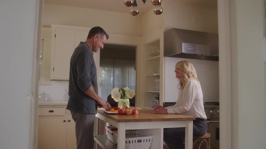 A man is cutting food while talking with a woman in the kitchen
