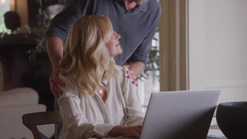 A man places his hands on the shoulders of a woman who is typing on a laptop and she turns her head to smile up at him
