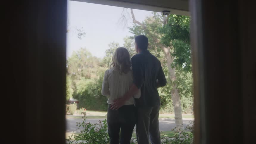 A couple stands arm in arm on their porch looking out at the quiet treelined street