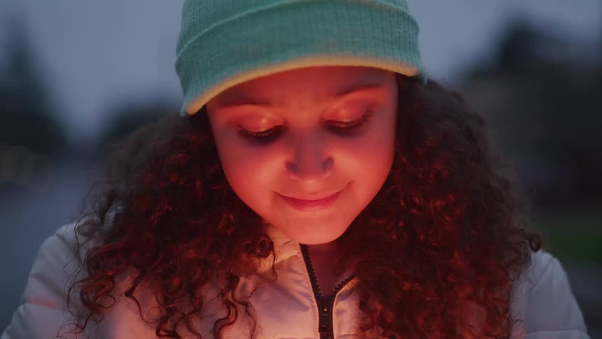 A young girl dressed in winter clothing stands beside a campfire with a smile on her face as the sun sets in the background