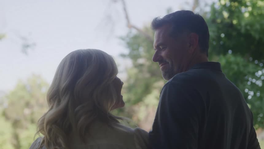 A middleaged couple with their backs to the camera turn to face one another with loving smiles in a bright sunlit outdoor setting