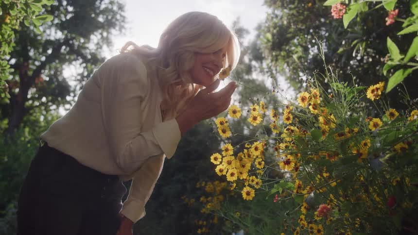 A woman with long blonde hair and a white shirt bends down to smell a bush of small yellow flowers in a sunny garden