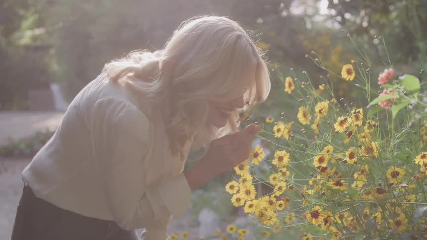 A woman with long blonde hair bathed in sunlight gently bends down to smell a cluster of yellow flowers in a garden