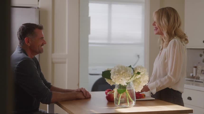 A woman chats with a seated man at a kitchen table both smiling warmly