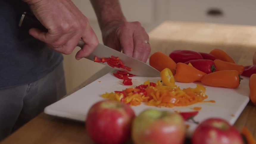 A closeup shot of a persons hands chopping red and orange bell peppers on a white cutting board with apples blurred in the foreground