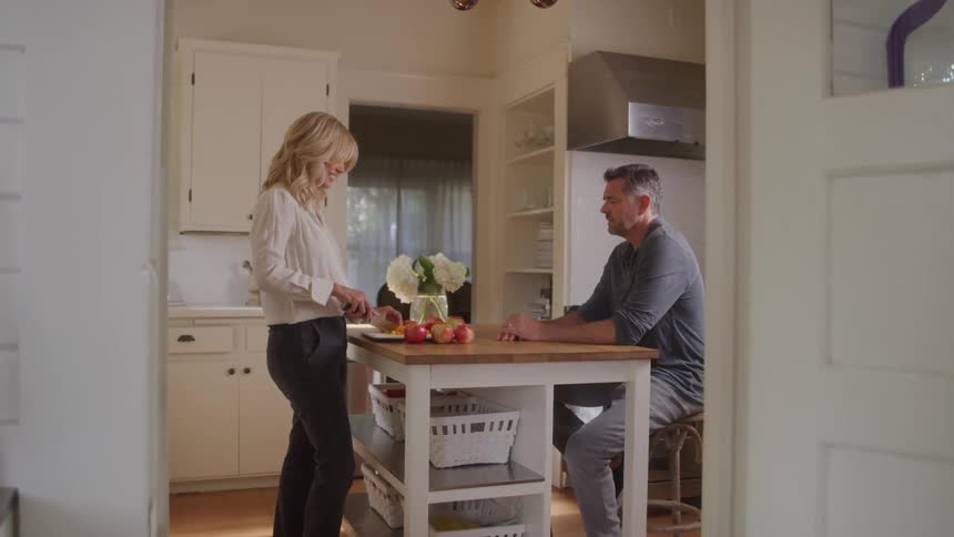A middleaged couple shares a quiet moment in their kitchen as the woman prepares food on a central island while the man watches