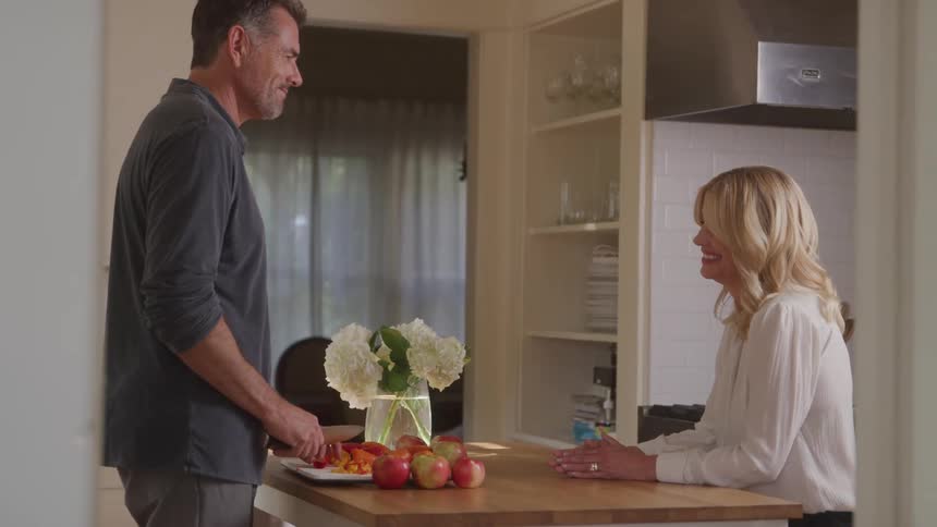 A man slices food at the kitchen island while a woman smiles at him from across the counter