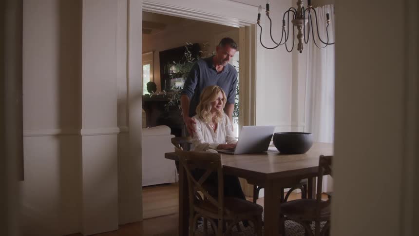 A man walks into the dining room as a woman works on her laptop at the tab