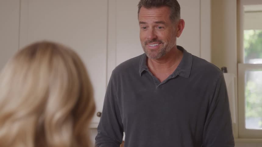 A man with short brown hair and a beard smiles and speaks to a woman with blonde hair before looking down while chopping vegetables on a cutting board in a kitchen