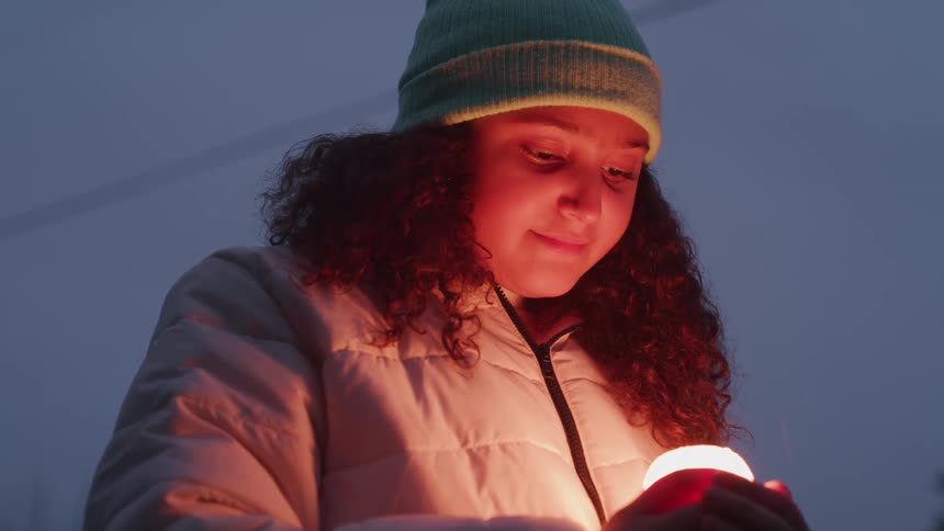 Girl in a light jacket and green beanie holds a lit candle at dusk