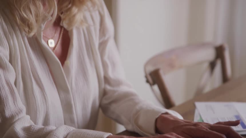 A woman with long blonde hair and a white collared shirt smiles while she types on a silver laptop