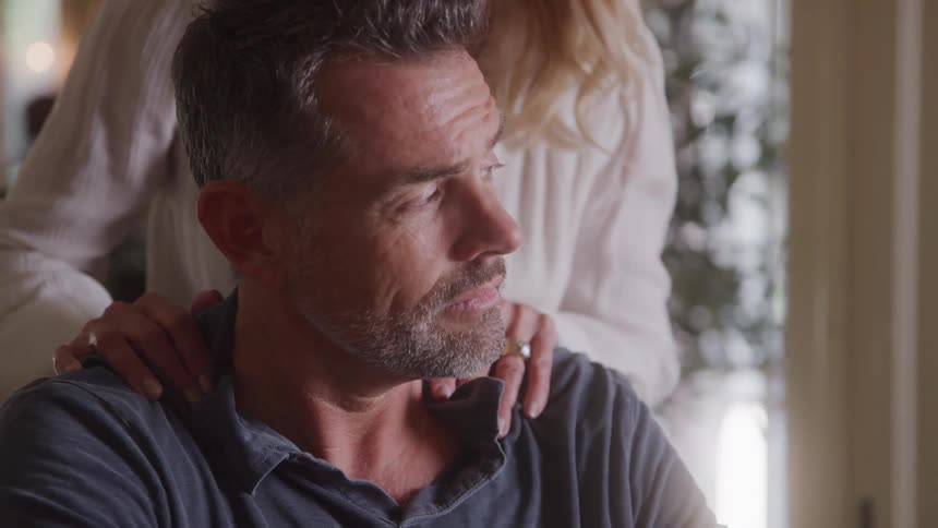 A loving middleaged woman embraces a man from behind offering comfort and support as he sits with a worried expression