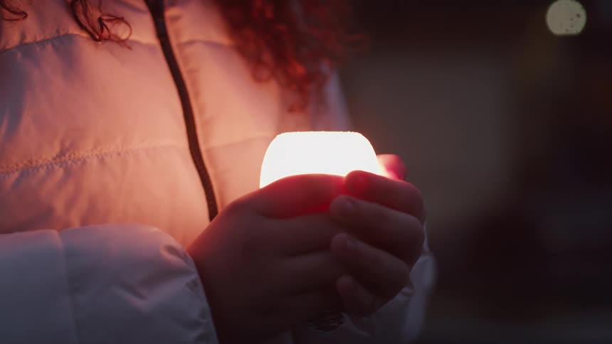 A girl with long wavy brown hair and a white puffer jacket holds a small glowing white lantern in both hands